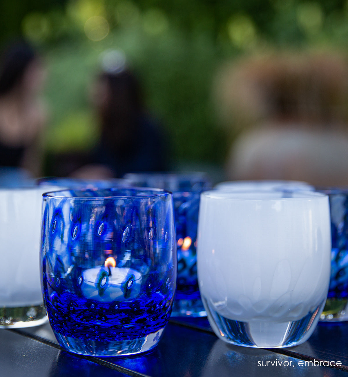 several survivor sapphire blue bubble, hand-blown glass votive candle holders on an outdoor table with embrace and friends sitting and chatting in blurred background.