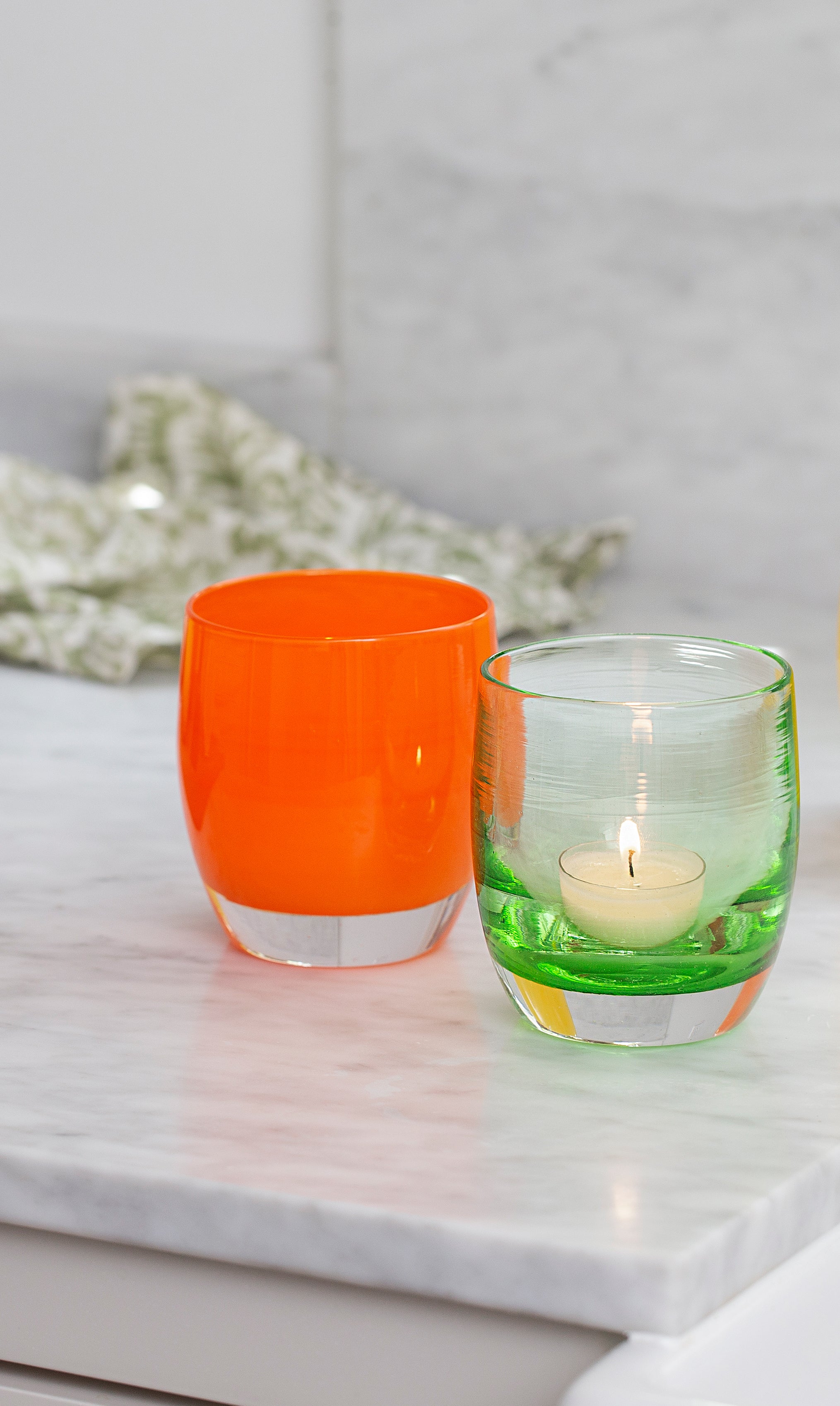 tangerine, bright orange hand crafted glass candle holder paired with clover, transparent green hand-blown glass candle holder on a marble table top.