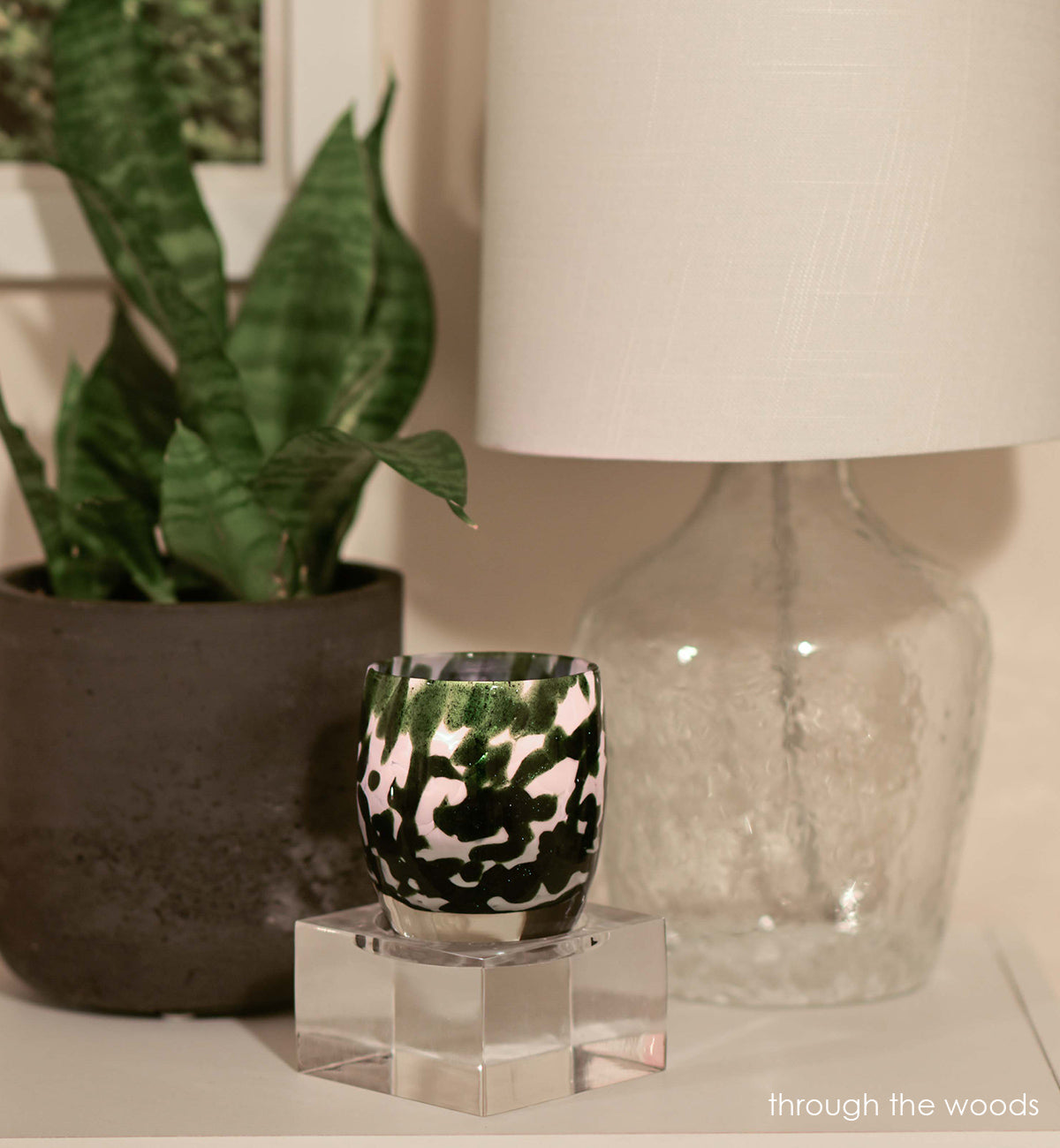 through the woods, white green texture, hand-blown-glass votive candle holder on a square baby stand next to a lamp and potted plant.