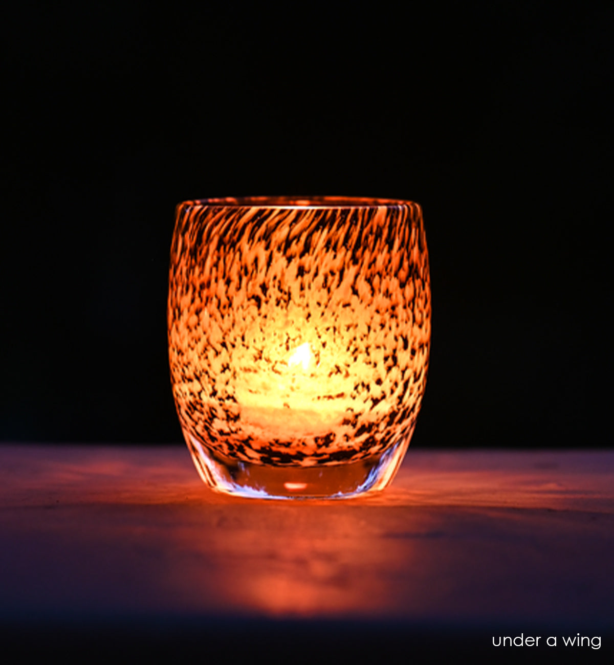 under a wing, white and brown feathered hand-blown glass votive candle holder shining light and shadows on a dark surface.