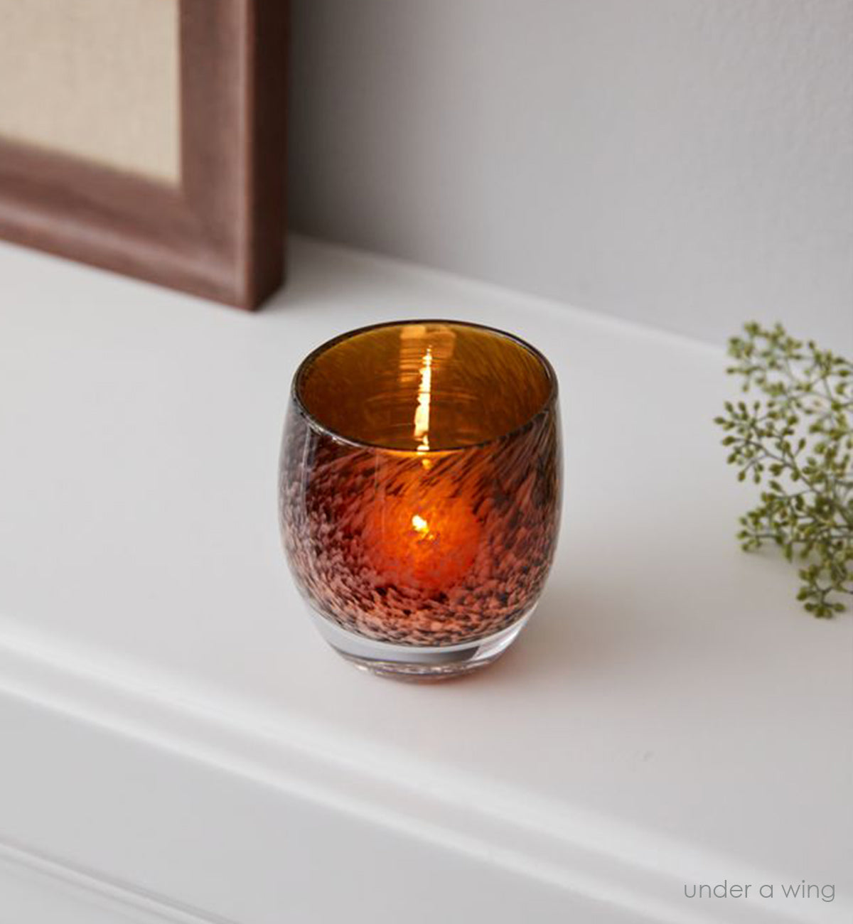 under a wing, an amber and brown mottled hand-blown glass votive candle holder on a white mantle with a plant and frame.