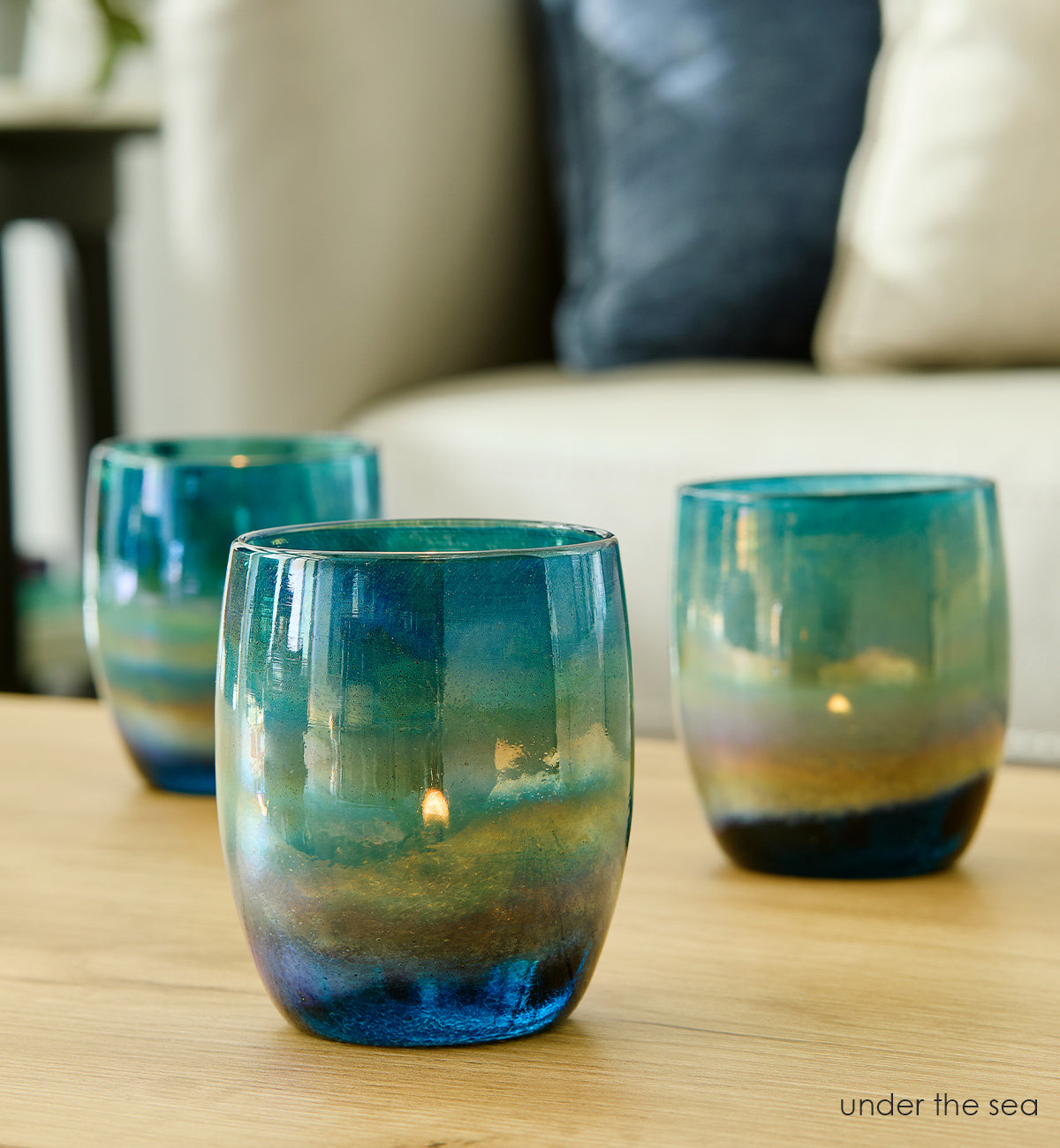 Three under the sea, blue green luster hand-blown glass votive candle holders with lit tealights and glowing flames inside placed on a light wood coffee table with cream couch in background.