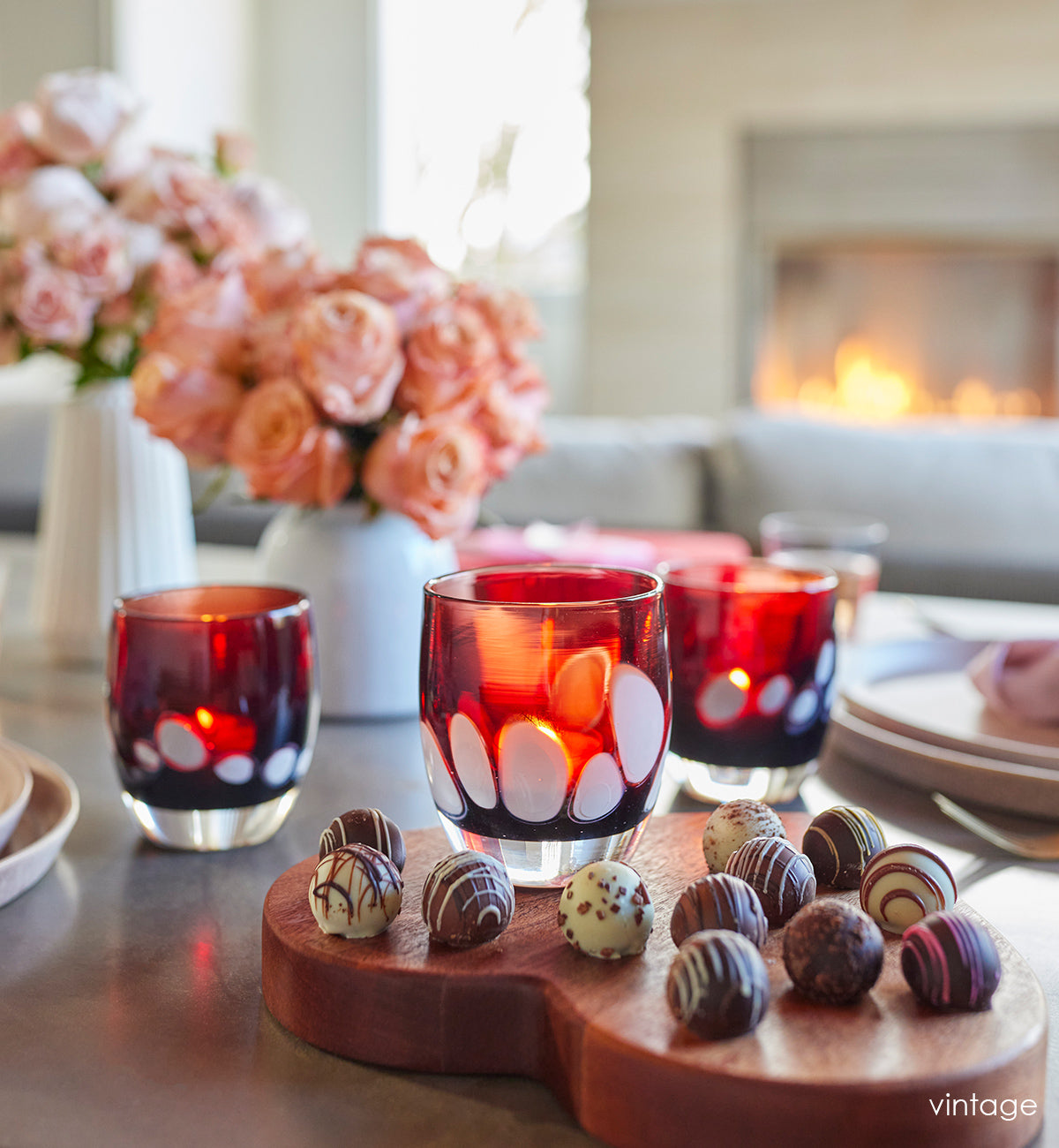 vintage red with white spots hand-blown glass candle holder with chocolates on wood heart and pink roses in white vase on a wood table.