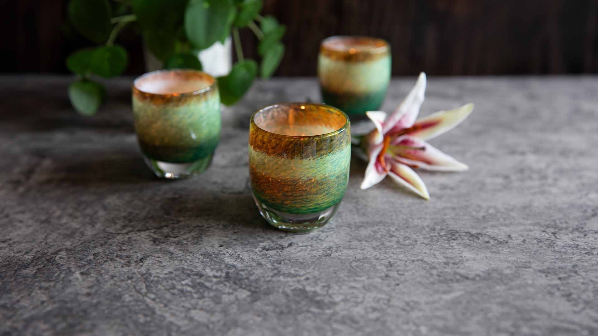 Three pond colored water lilies votive candle holders lit on a stone table with a pilea plant and a lily flower.
