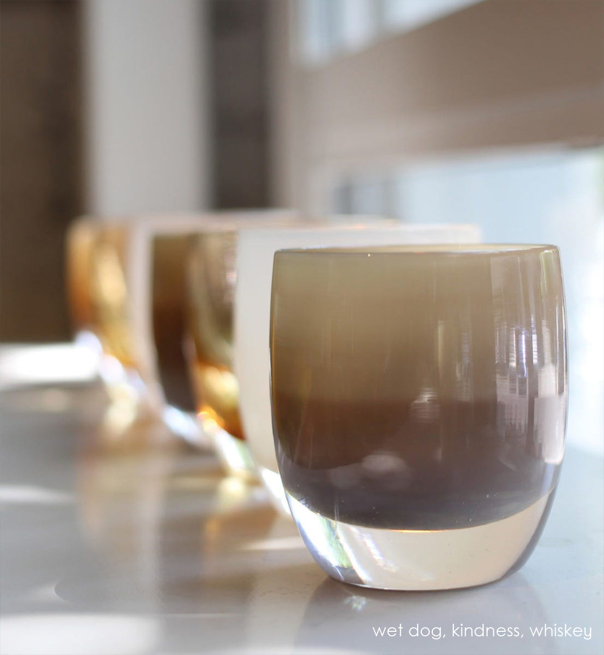 wet dog, two toned brown, hand-blown glass votive candle holder in a line of votives including whiskey and kindness in front of a window.