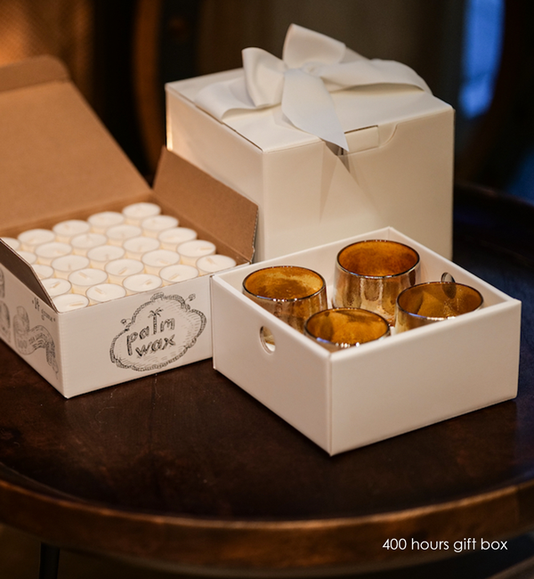 four hundred hours gift box, one hundred palm wax tea lights, and four hand-blown glass votives candle holders with a bowed white box on a dark wood table.