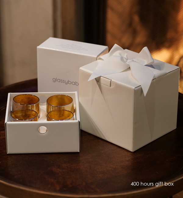 400 hours gift box; 100 palm wax tealights with four stay gold hand-blown glass votive candle holders and bowed white box on a dark circular side table.