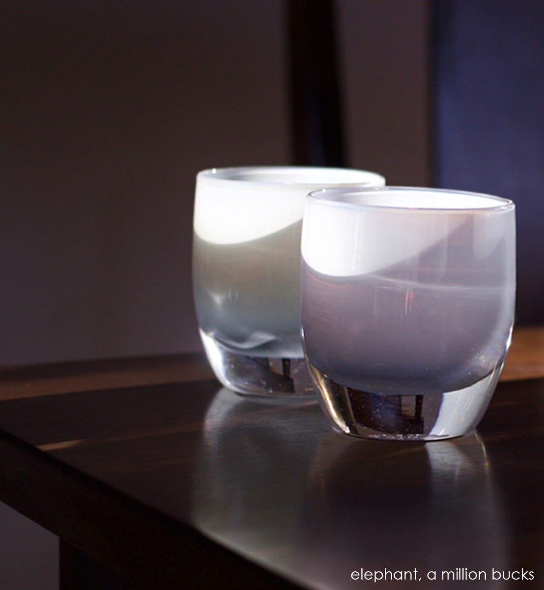 a million bucks light grey hand-blown glass votive candle holder. Paired with elephant sitting in sunlight on dark wood table.
