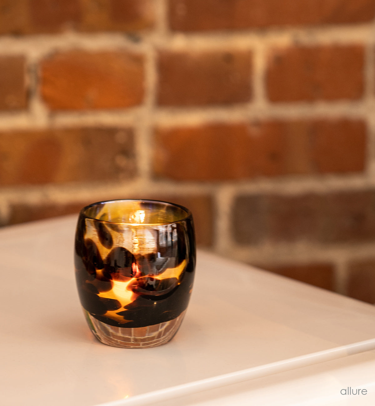 allure brown and gold hand-blown glass votive candle holder on a white dresser in front of a brick wall.