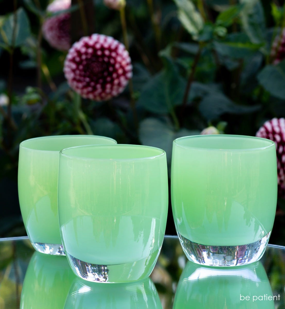three be patient, light green hand-blown glass votive candle holders on a mirror table outside with flowers behind.