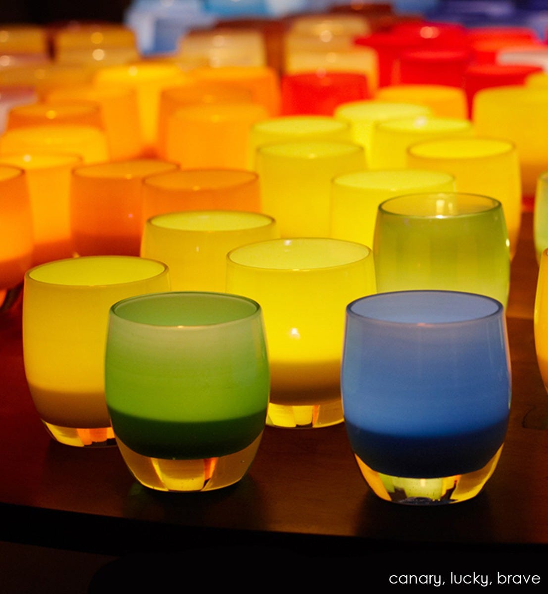 brave egyptian blue hand-blown glass votive candle holder grouped with lucky and canary, a lit on a dark wood table.