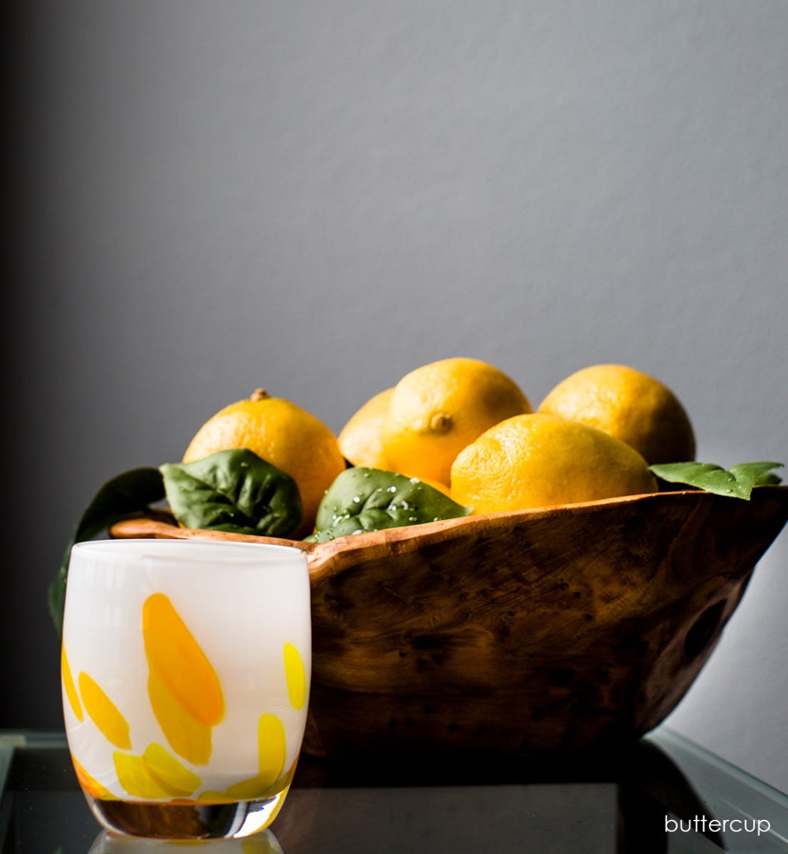 buttercup, yellow petals on white, hand-blown glass votive candle holder on a glass table with a bowl of lemons behind it.