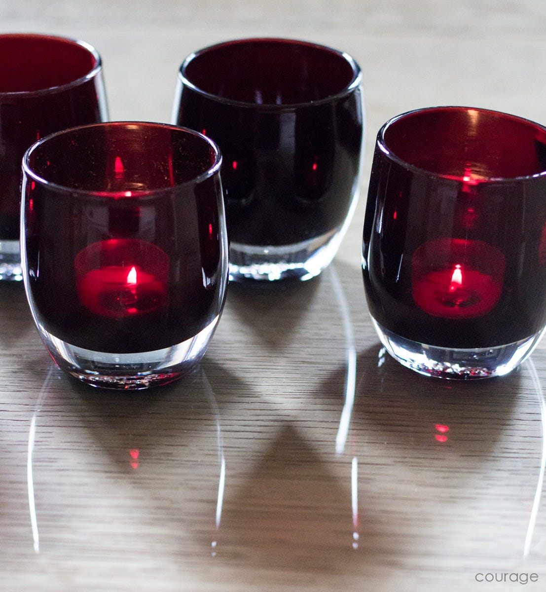 several courage, translucent mahogany red, hand-blown glass votive candle holders lit with tealights on a light wood surface.