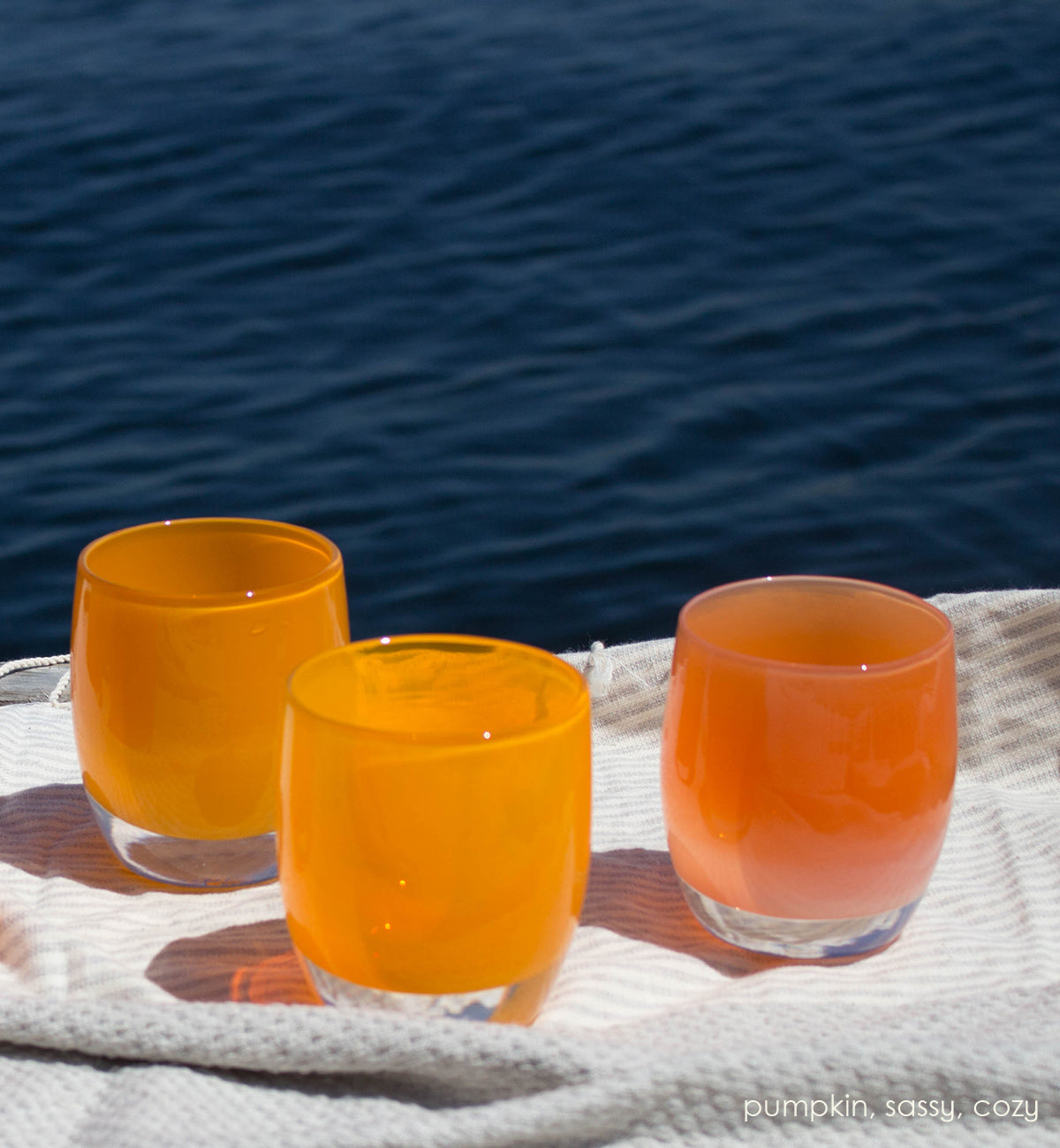 cozy, stripey apricot orange hand-blown glass votive candle holder with sassy and cozy on a beach towel in front of a body of water.