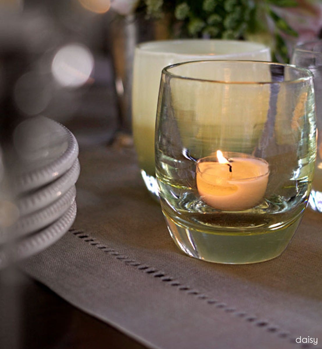 daisy transparent crystal yellow hand-blown glass votive candle holder placed on a table runner on a dining room table next to ceramic plates.