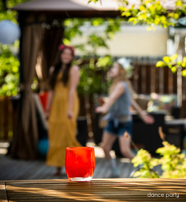 dance party ginger red, hand-blown glass votive candle holder placed on patio table, with two girls in crowns have a dancing party of their own in background.