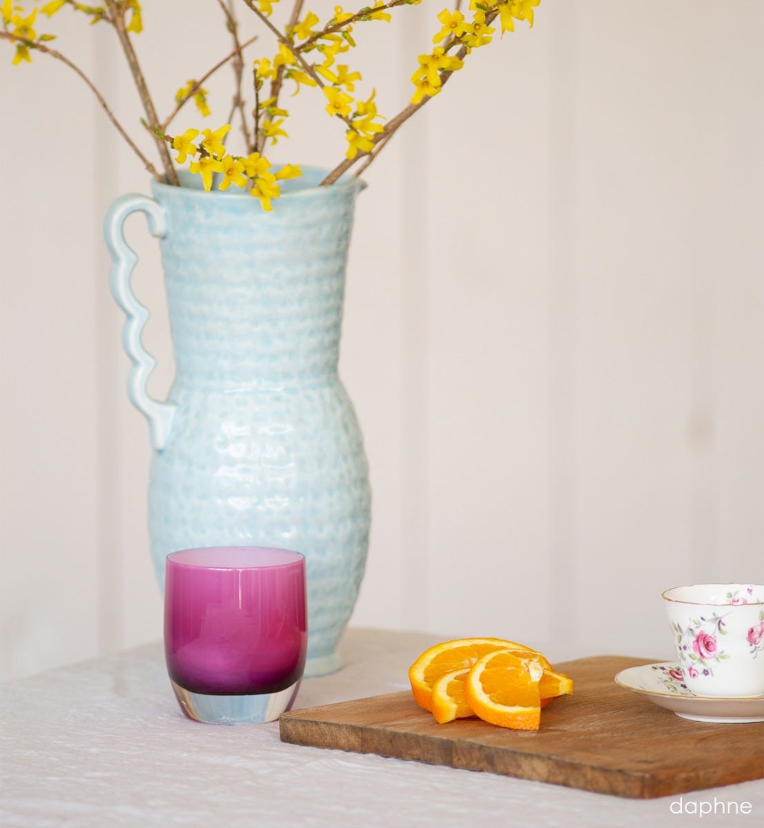 daphne bright purple with white interior, hand-blown glass votive candle holder on a table with a light bue vase, and tray holding sliced oranges and a teacup.