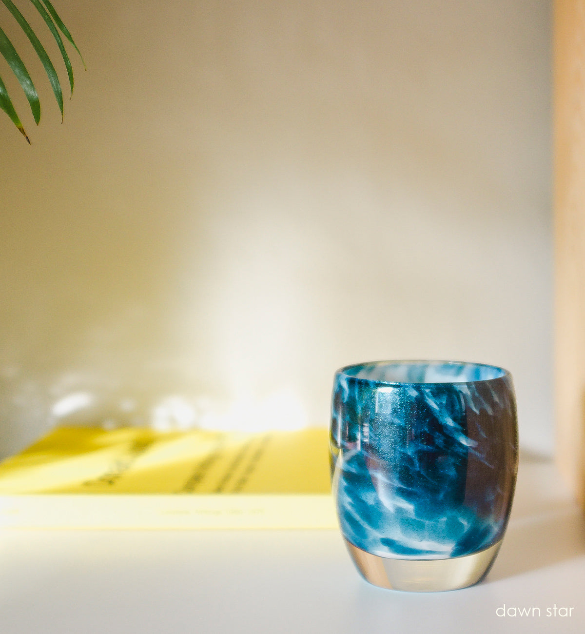 dawn star blue glitter, hand-blown glass votive candle holder sitting on a cream surface with a yellow coffee table book behind.