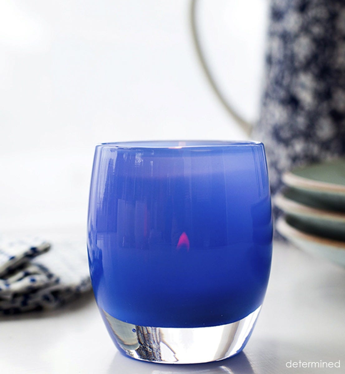 determined sapphire blue hand-blown glass votive candle holder with patterned napkins and plates blurred in background.