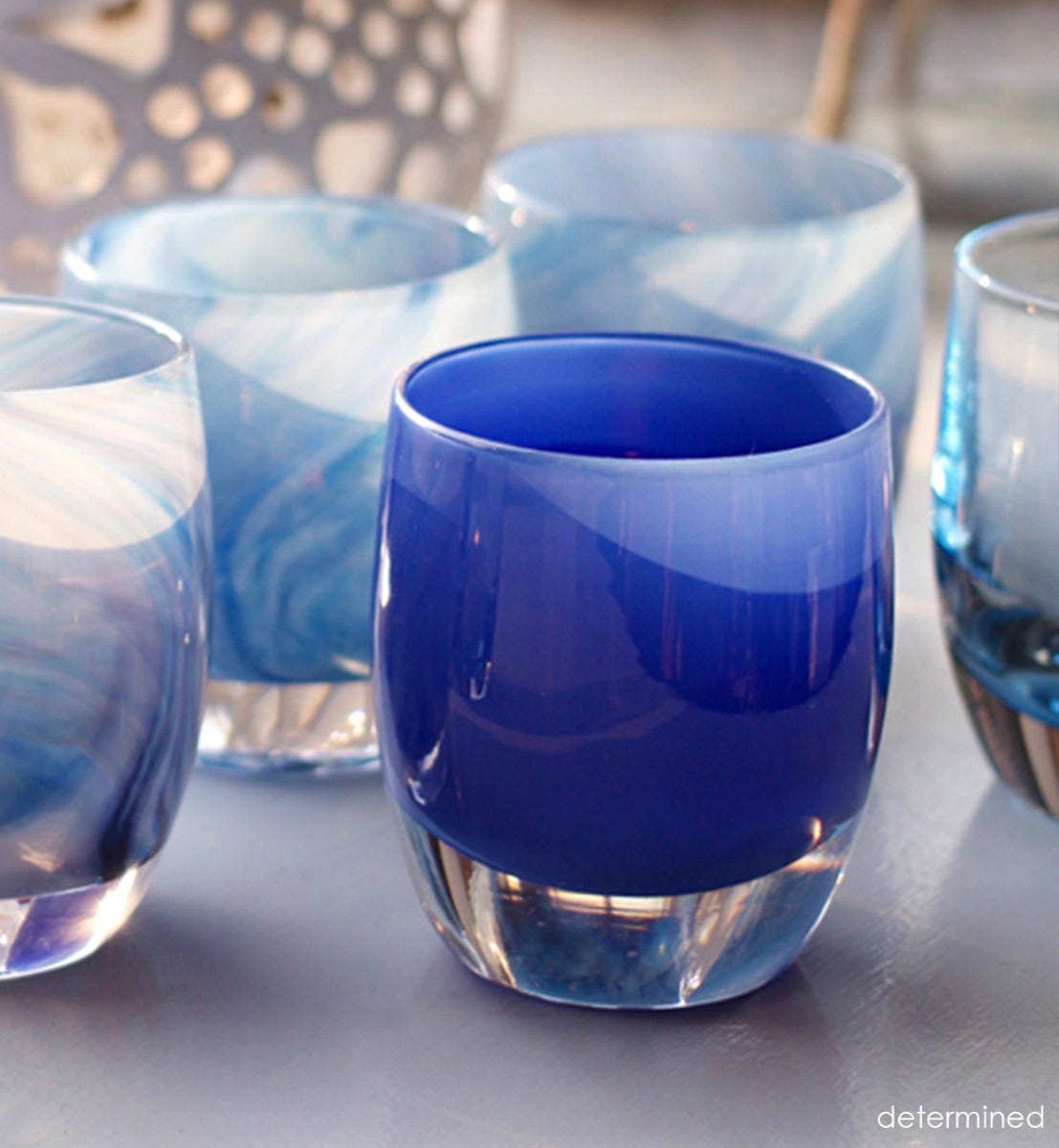 determined sapphire blue hand-blown glass votive candle holder with other votives with a ceramic patterned lamp in background.