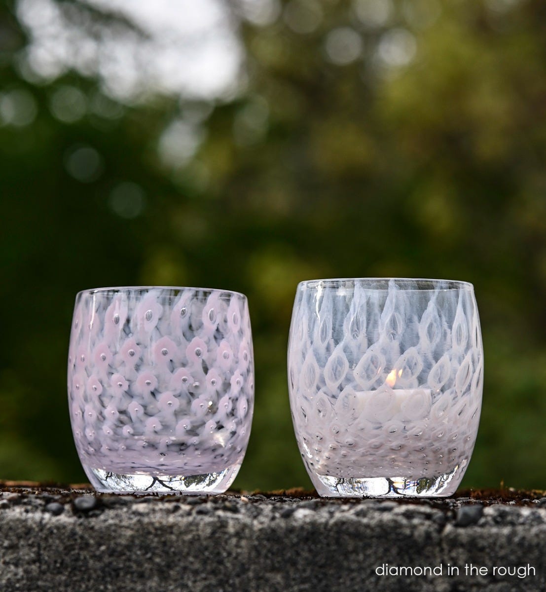two diamond in the rough, light pink with bubble pattern hand-blown glass votive candle holders sitting on concrete wall with blurred trees in background.