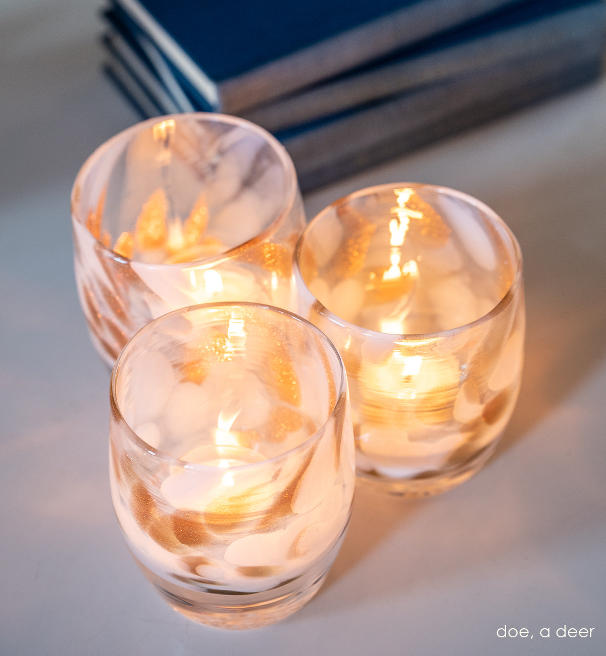 three doe, a deer shimmering gold and white petal, hand-blown glass votive candle holders on a white surface next to a stack of blue books.