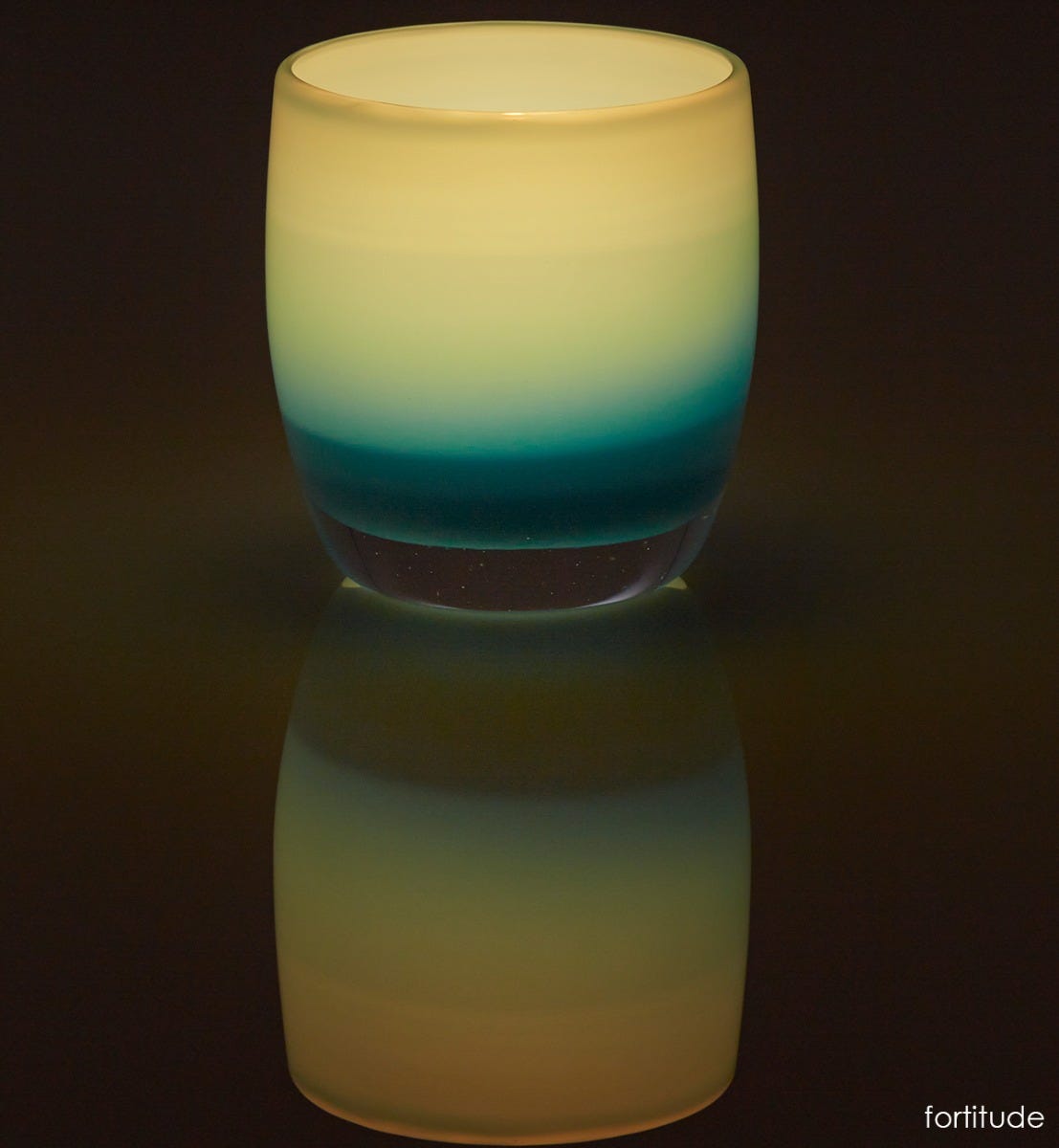 fortitude carolina blue hand-blown glass votive candle holder lit on a reflective surface.