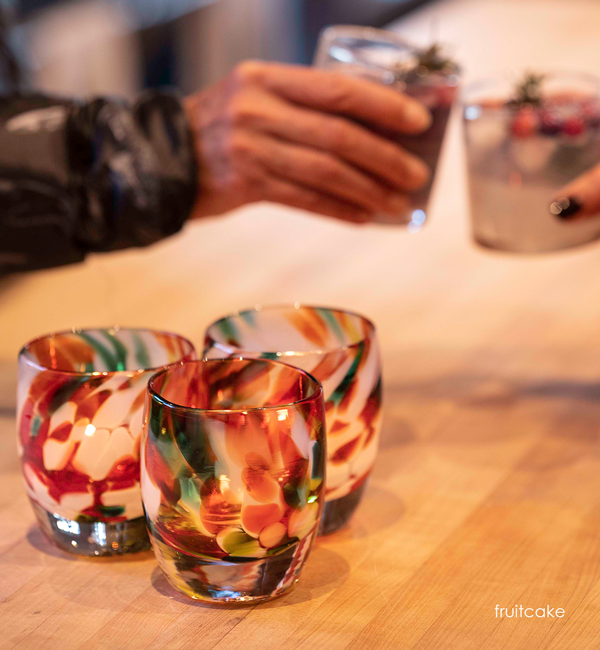a few fruitcake, multicolored petal texture, hand-blown glass votive candle holders on a light wood kitchen counter with two people toasting their filled rockers in background.