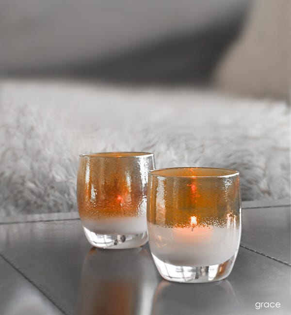 two grace golden topped white hand-blown glass votive candle holders on a metal table with furry blanket behind.