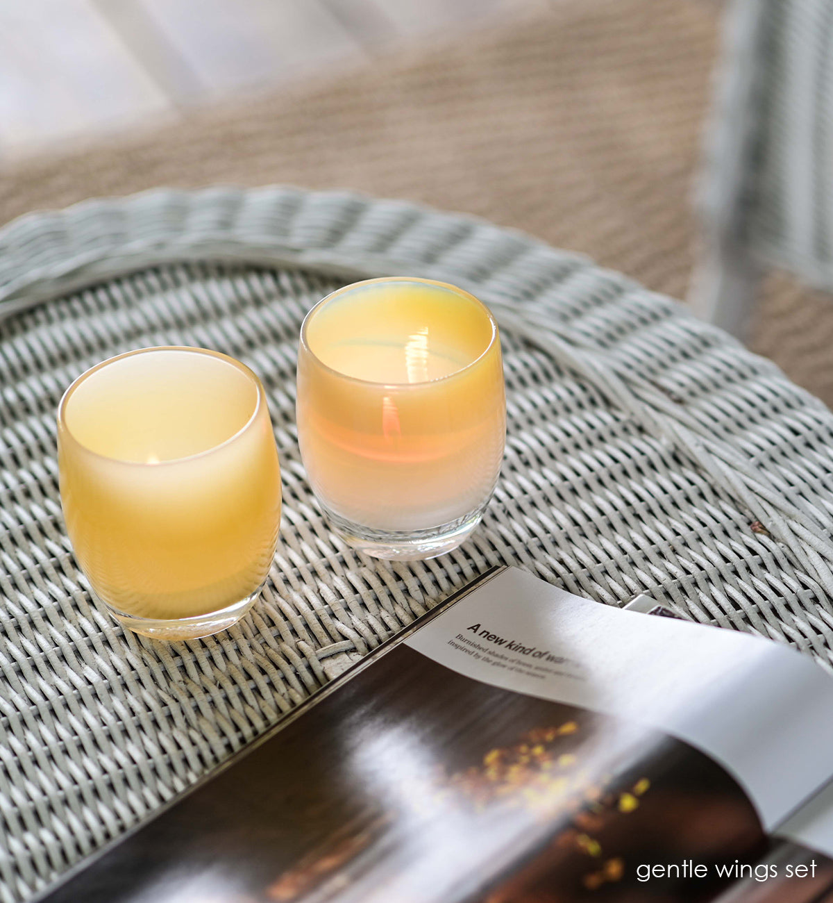 gentle wings set, angel and comfort hand-blown pale yellow set of two, hand-blown glass votive candle holders next to a magazine on a wicker coffee table.