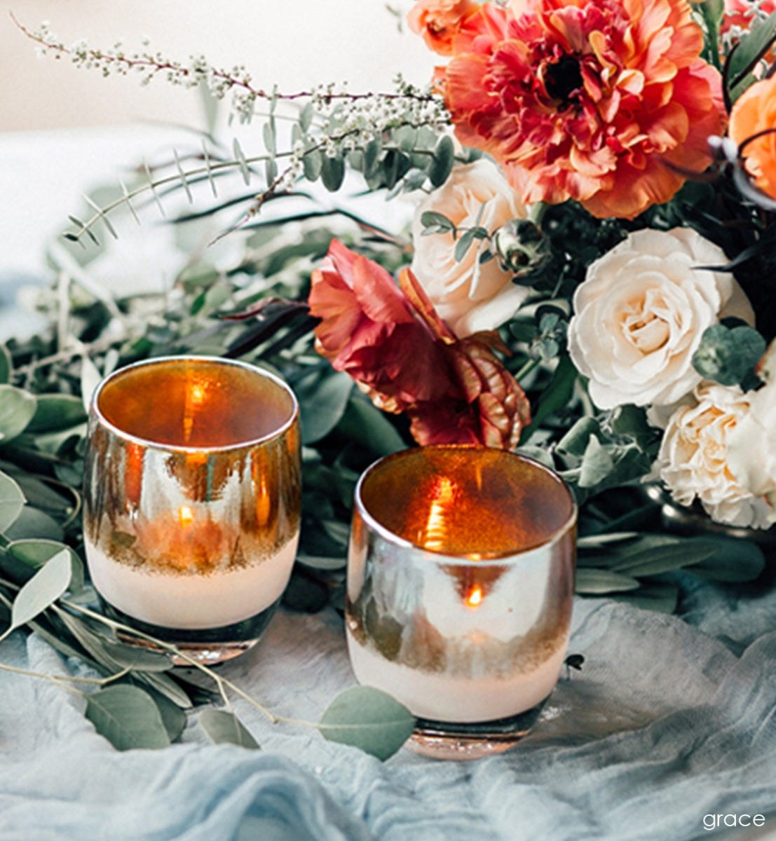 two grace golden topped white hand-blown glass votive candle holders next to a bouquet of flowers.