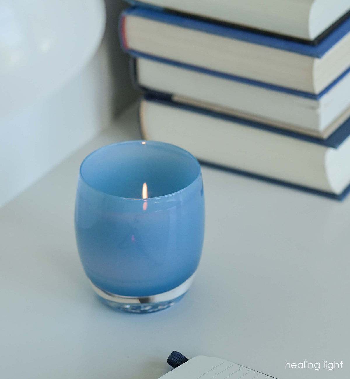 healing light sky blue hand-blown glass votive candle holder next to a journal and stack of books on a white surface.