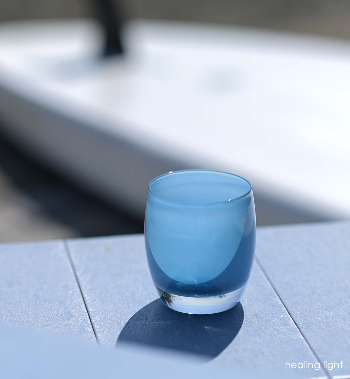 healing light sky blue hand-blown glass votive candle holder sitting on a blue patio in the sunlight.