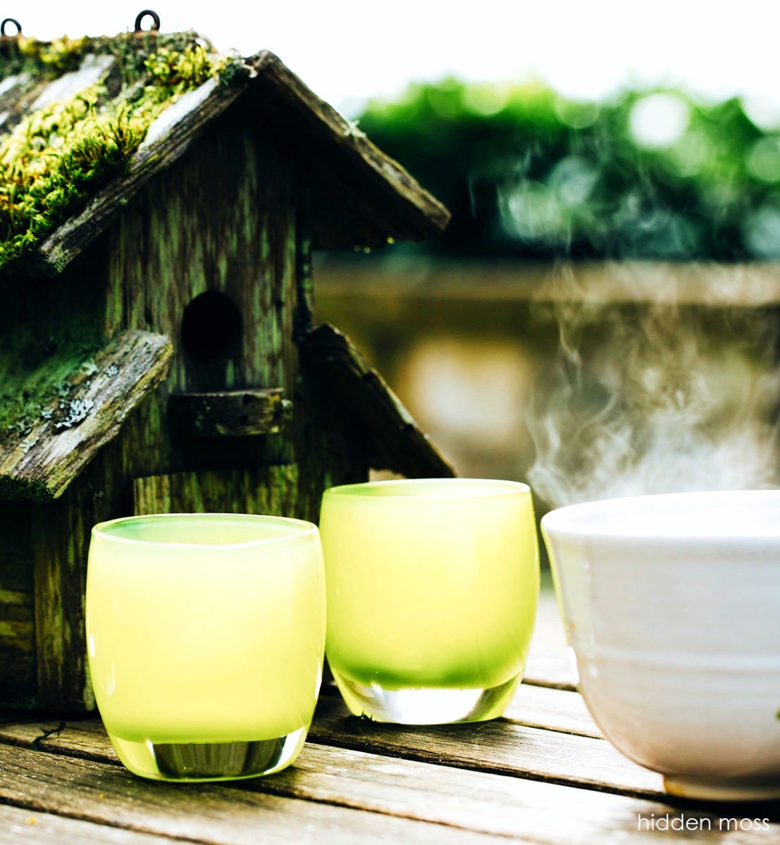 hidden moss, pear green, hand-blown glass votive candle holder next to bowl of steaming contents and mossy birdhouse.