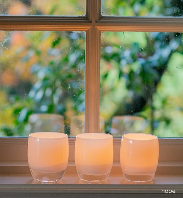 three lit hope pure white hand-blown glass votive candle holders on a white windowsill with greenery outside.