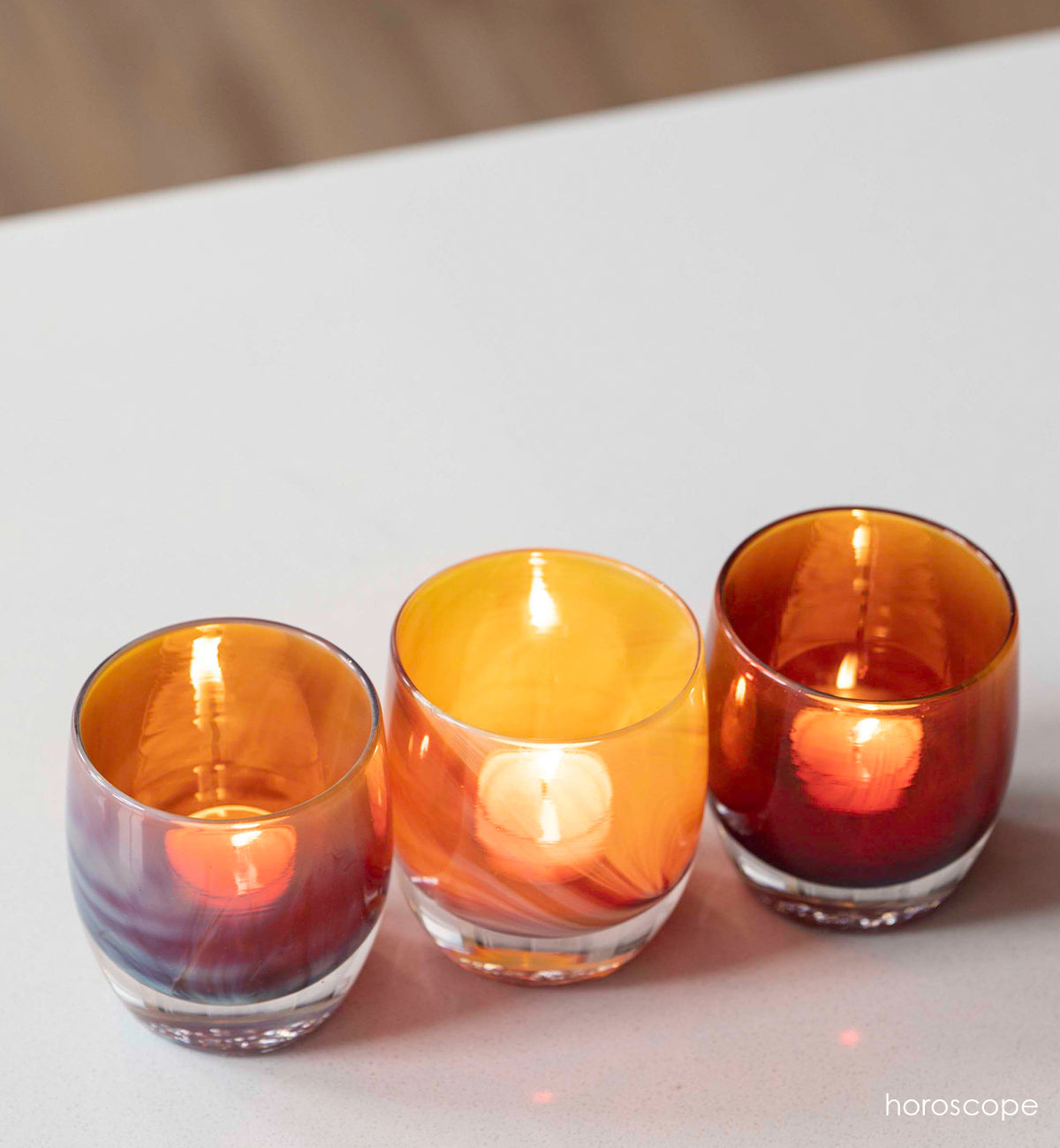 three horoscope purple orange, hand-blown glass votive candle holders lit on a white table.