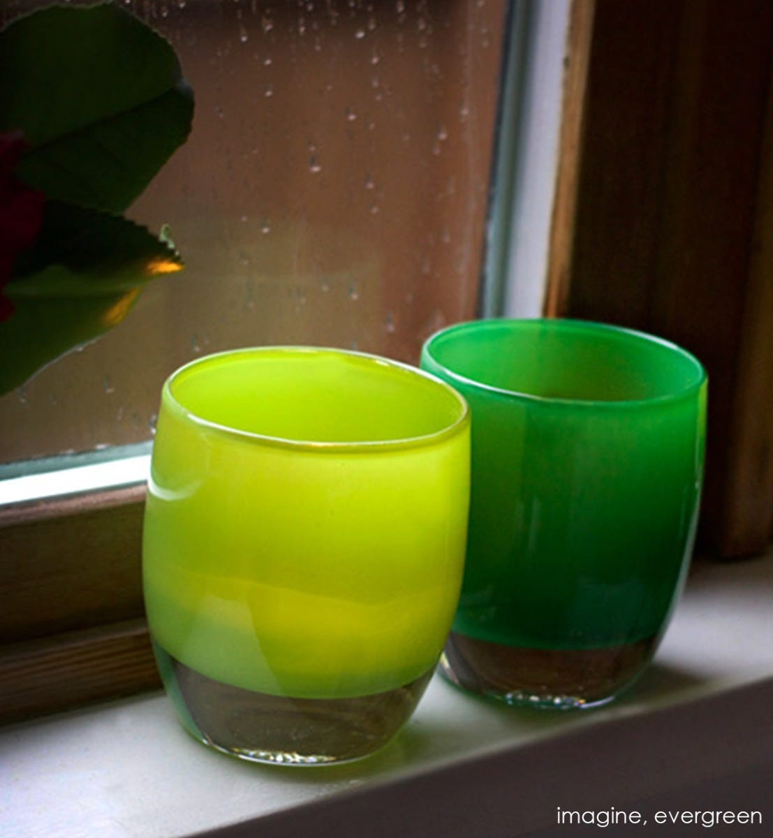 imagine, kashmir green, hand-blown glass votive candle holder. Paired with evergreen sitting on a cream windowsill with rain outside.