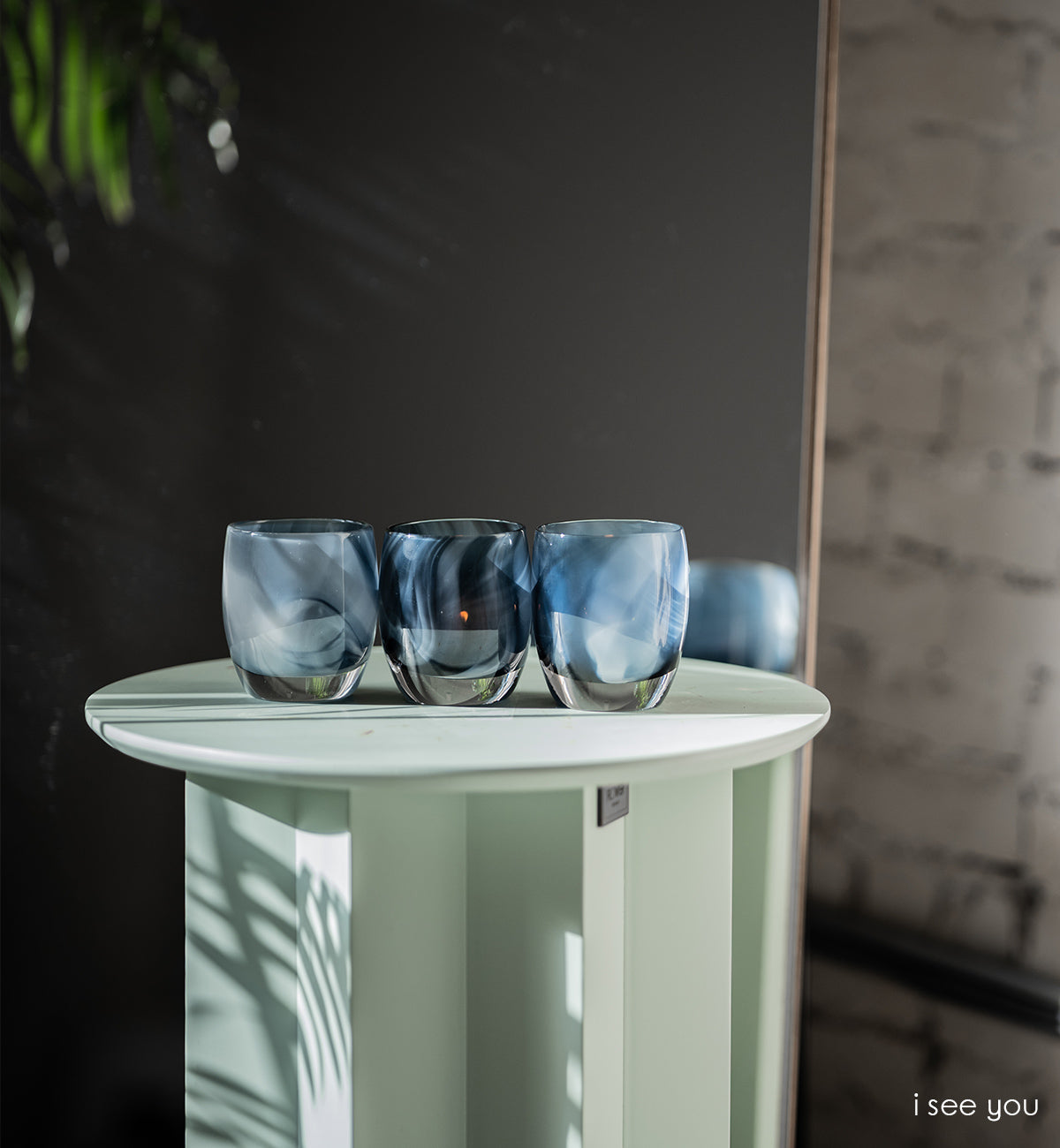 three i see you, smokey blue pattern, hand-blown glass votive candle holders side by side on a white table next to a mirror.