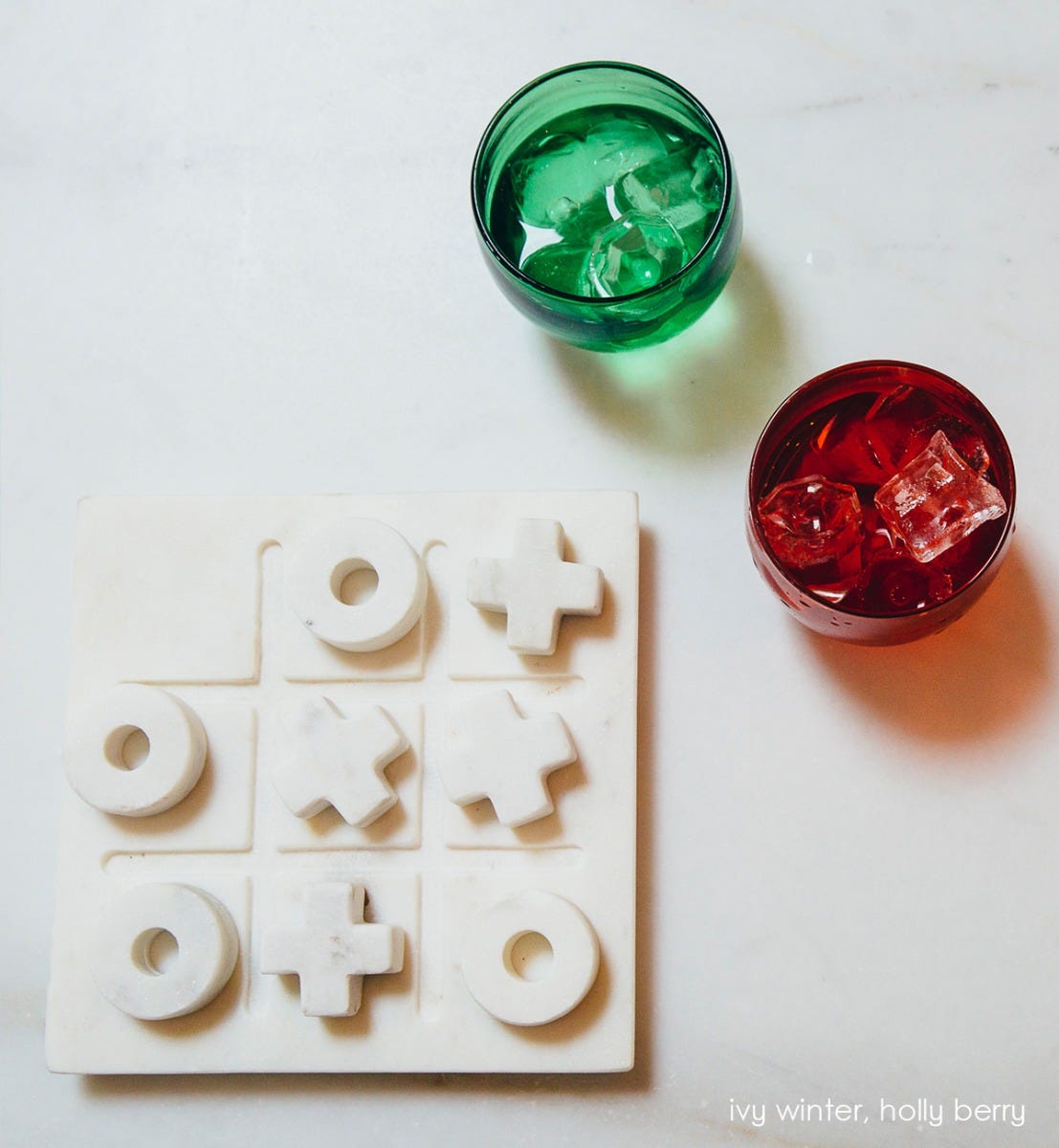 ivy winter drinker transparent green hand-blown drinking glasswith holly berry drinker, both filled with ice and liquid next to a tic tac toe set.