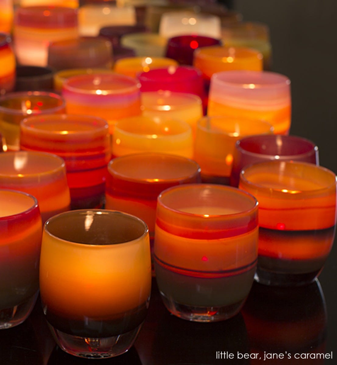 several jane's caramel, caramel brown and purple gradient, hand-blown glass votive candle holders all lit with tealights in a glassybaby display with little bear.