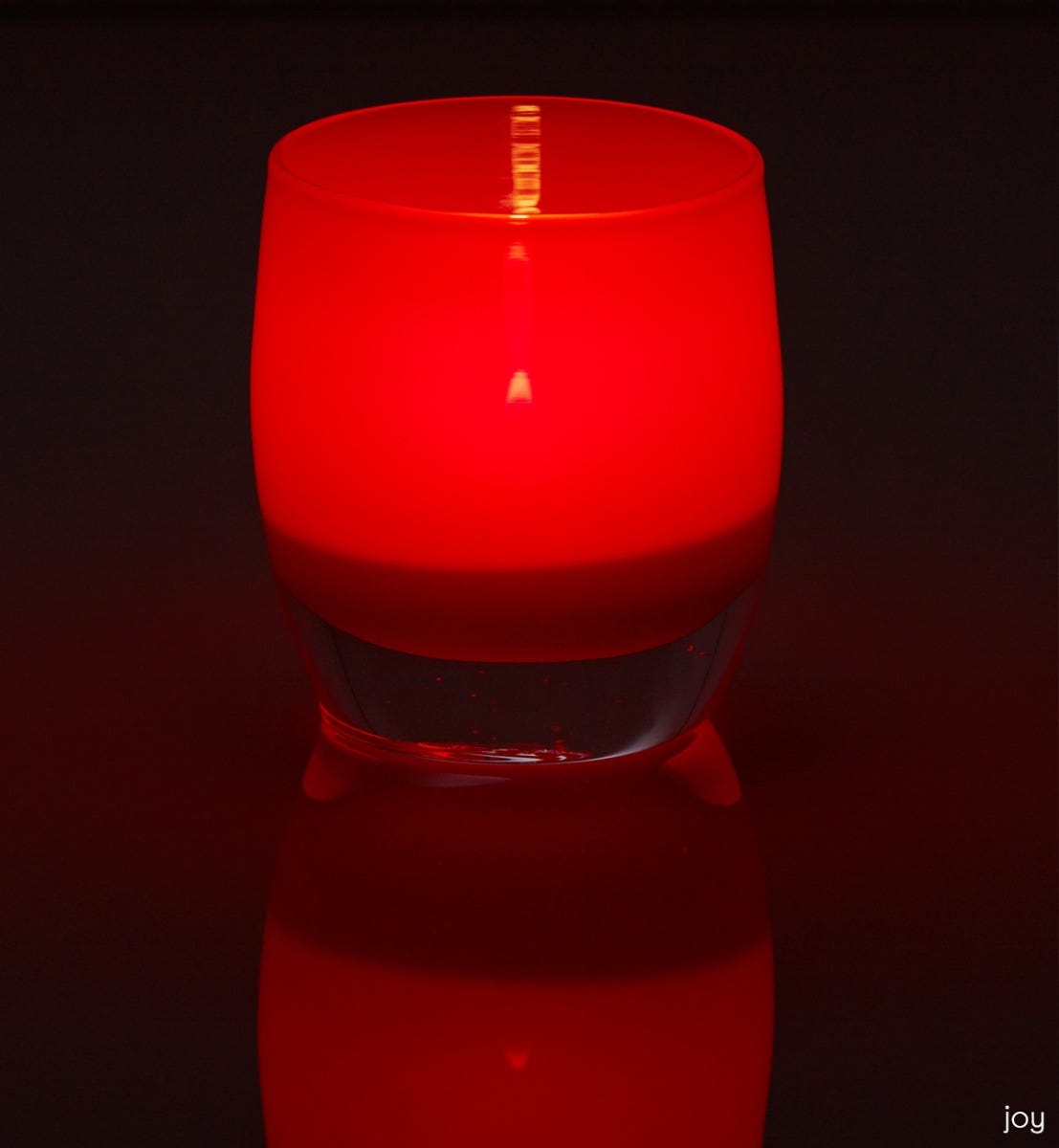 joy crimson red hand-blown glass votive candle holder on a reflective surface.