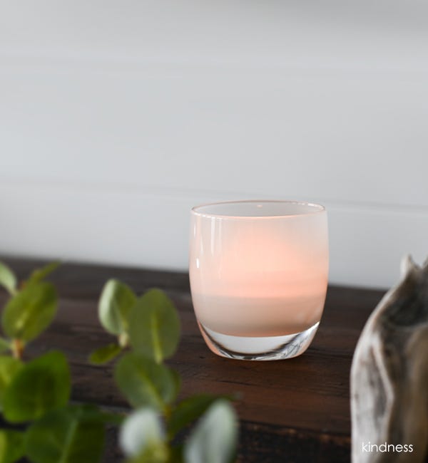 kindness soft cream hand-blown glass votive candle holder on a wood surface with white background and plant.