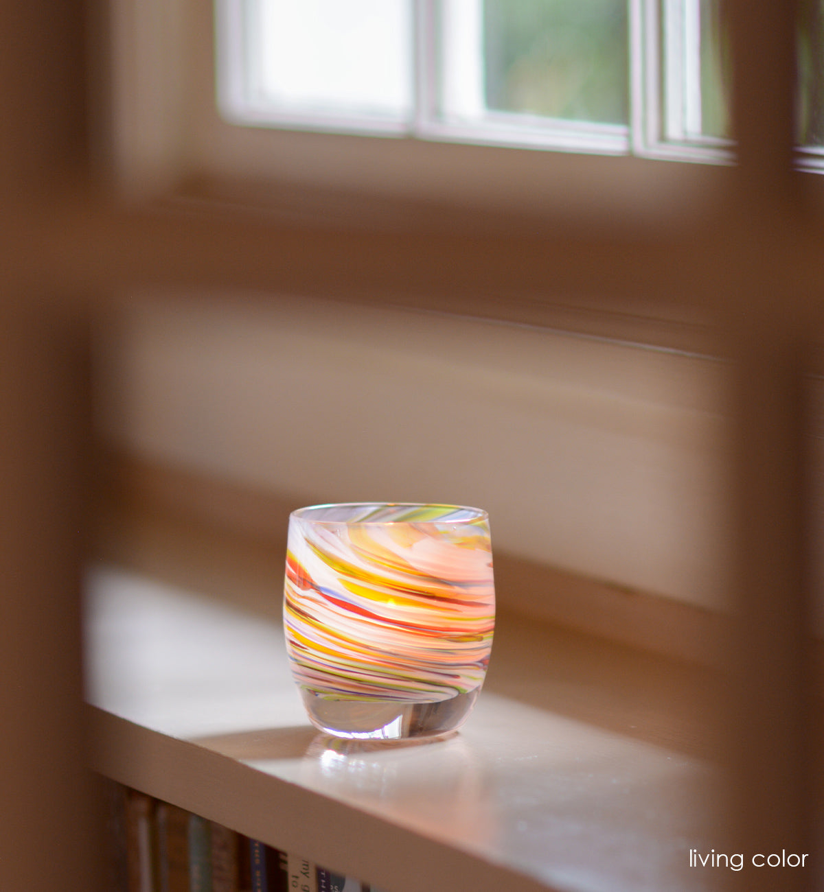 living color hand-blown multicolored swirl, hand-blown glass votive candle holder on a cream mantle beneath a window.