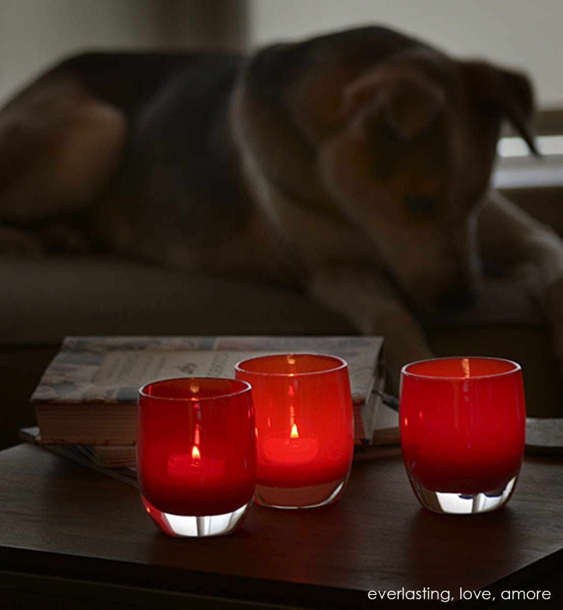 love brick red hand-blown glass votive candle holder. Paired with everlasting and amore on a coffee table with books and a dog in the background.