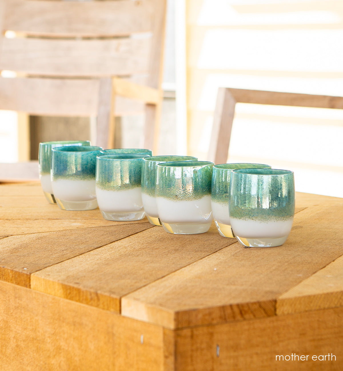 several mother earth white with metallic green blue top, hand-blown glass votive candle holders on a light wood table outside.