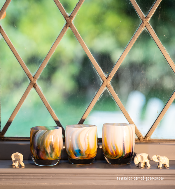 three music and peace, rainbow swirled texture, hand-blown glass votive candle holders next to ceramic elephants in front of a metal framed window.