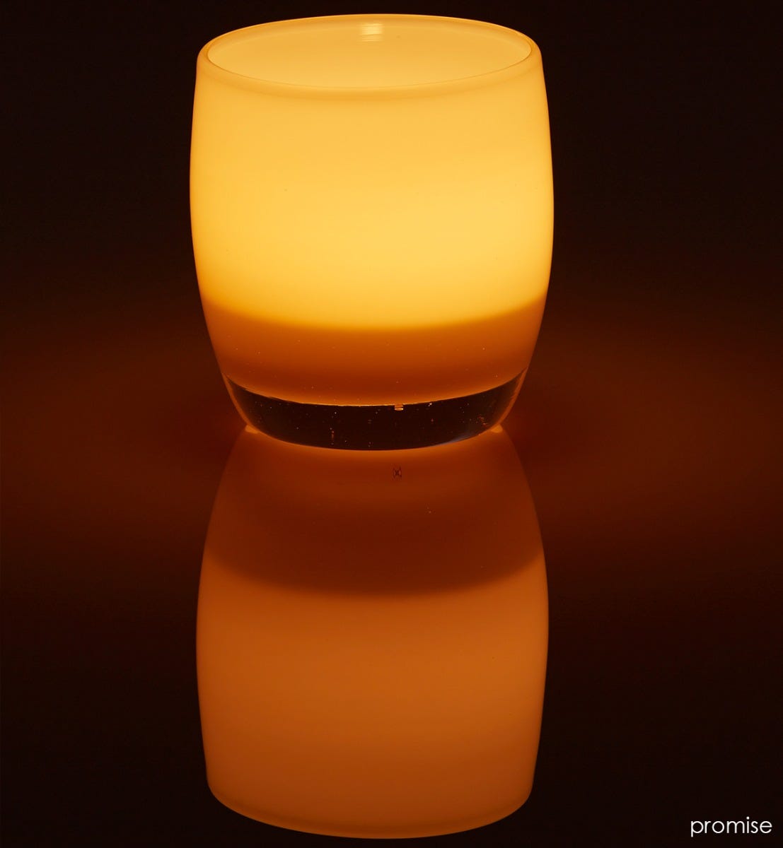 promise cream hand-blown glass votive candle holder lit on a dark reflective surface.