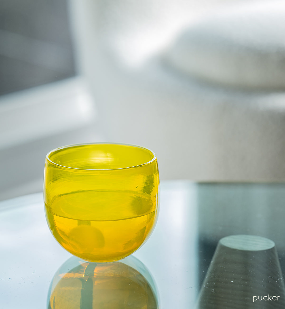 pucker drinker, bright yellow hand-blown drinking glass with and ice cube and liquid on a reflective glass coffee table.