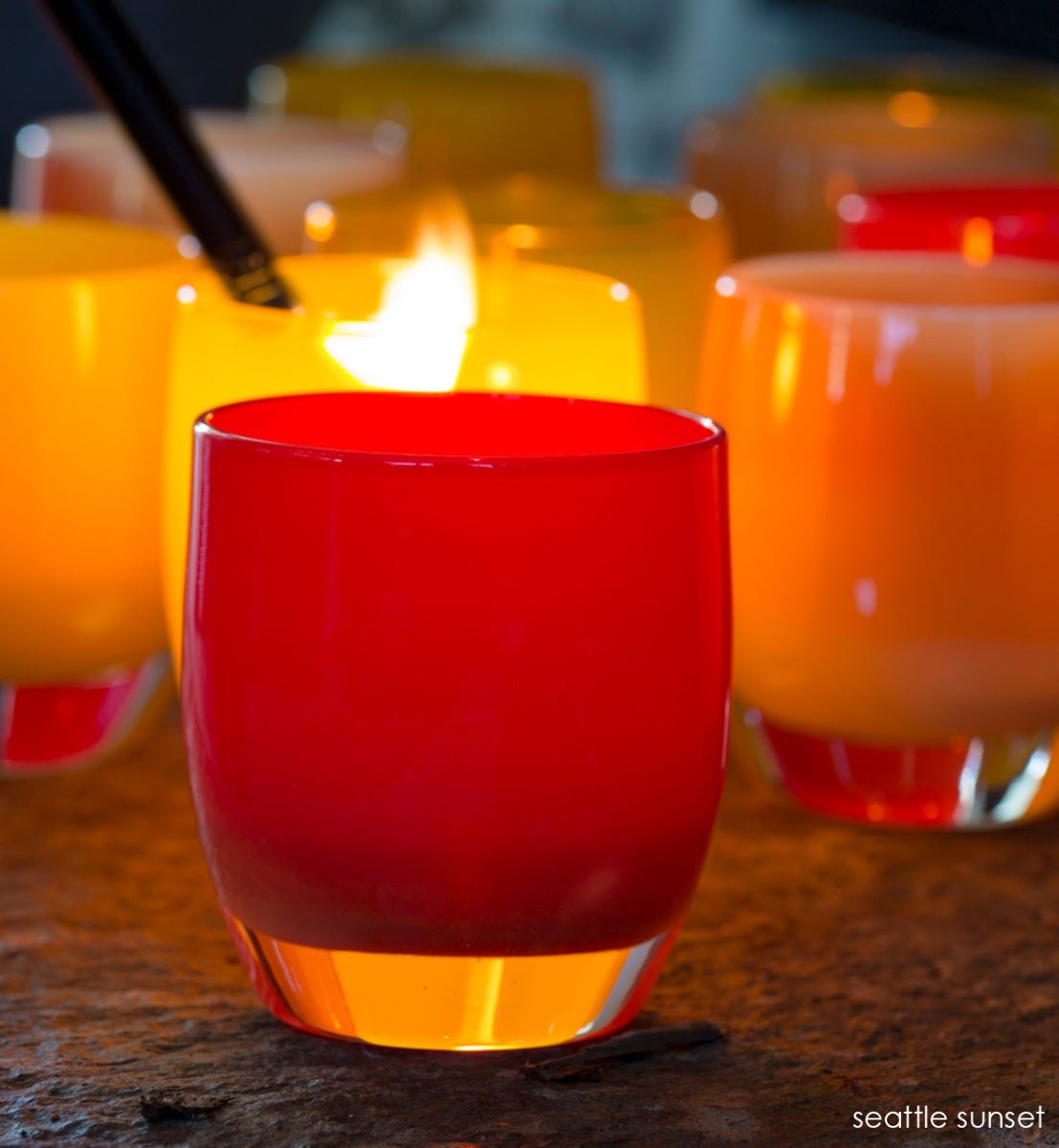 seattle sunset, bright orange red, hand-blown glass votive candle holder on a rocky material table with other votives being lit in background.