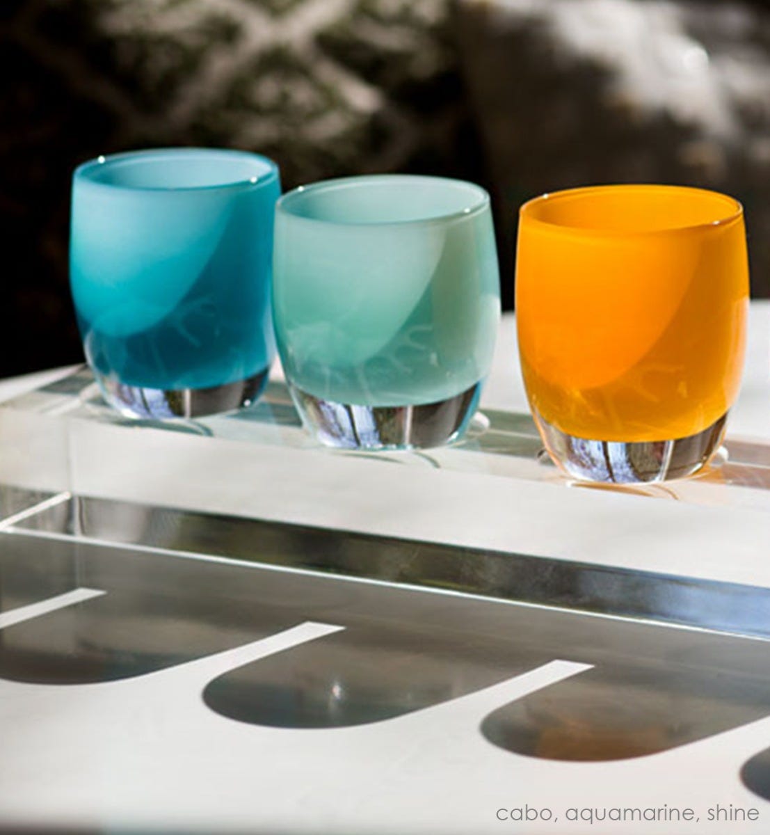 shine hand-blown bright orange glass votive candle holder with aquamarine and cabo on an acrylic stand sitting in the sun.